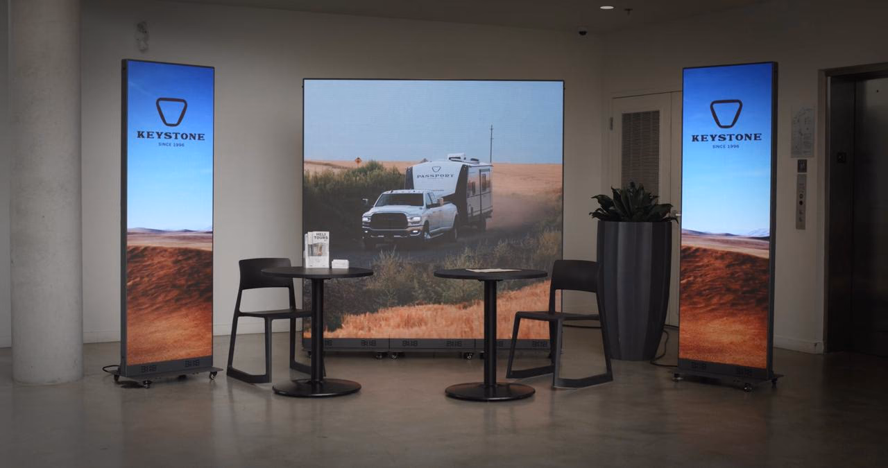 Keystone RV lounge: a video wall flanked by two LED totems, with cafΓ© tables in front